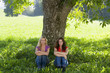 Two women sitting apart, leaning against a tree.