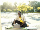 Photo: Male university student sitting on ground with textbooks, using laptop, smiling, portrait Male university student sitting on ground with textbooks, using laptop, smiling, portrait