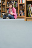 Photo: Girl (10-12) in spectacles sitting on floor beside bookshelf in library, reading book, surface level Girl (10-12) in spectacles sitting on floor beside bookshelf in library, reading book, surface level