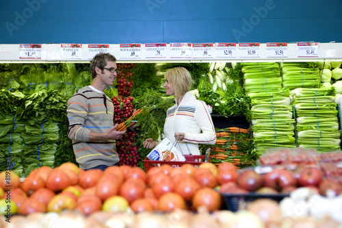 Photo: Couple shopping in vegetable section of supermarket, smiling, side view Couple shopping in vegetable section of supermarket, smiling, side view