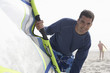 Photo: Man in wetsuit holding windsurfing board on beach, smiling, portrait, woman in background Man in wetsuit holding windsurfing board on beach, smiling, portrait, woman in background