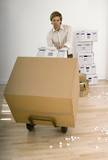Photo: Businessman moving large cardboard box with hand truck in sparse office, packing foam on floor Businessman moving large cardboard box with hand truck in sparse office, packing foam on floor