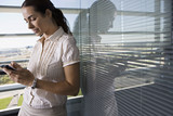Photo: Businesswoman leaning against office window, using personal electronic organiser, side view Businesswoman leaning against office window, using personal electronic organiser, side view