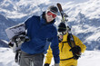 Male skier and male snowboarder walking up mountain ridge carrying skis