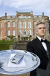 Mature butler with mobile phone on tray by manor house, low angle view