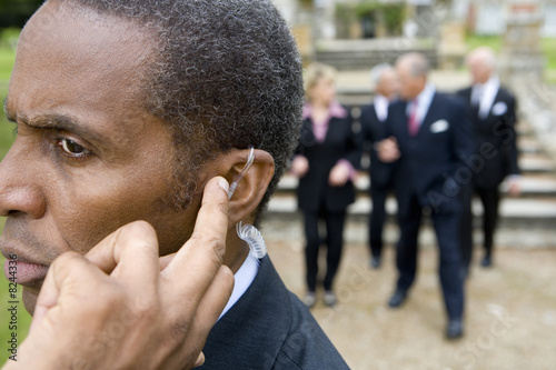 Photo: Man with hand on earpiece by manor house, businessmen and women in background, close-up Man with hand on earpiece by manor house, businessmen and women in background, close-up