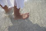 Photo: Woman standing on beach, ankle deep in sea water, low section, focus on feet, close-up, side view Woman standing on beach, ankle deep in sea water, low section, focus on feet, close-up, side view