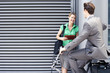 Photo: Businessman cycling on folding commuter bicycle flirting with businesswoman standing on pavement Businessman cycling on folding commuter bicycle flirting with businesswoman standing on pavement