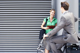 Photo: Businessman cycling on folding commuter bicycle flirting with businesswoman standing on pavement Businessman cycling on folding commuter bicycle flirting with businesswoman standing on pavement