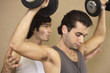 Photo: Close-up of a young man exercising with dumbbells and his instructor supporting him from behind Close-up of a young man exercising with dumbbells and his instructor supporting him from behind