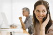 Photo: Businesswoman wearing a headset with a businessman working on a computer in the background Businesswoman wearing a headset with a businessman working on a computer in the background