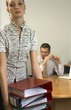Photo: Portrait of a businesswoman lifting a stack of files with a businessman sitting in the background Portrait of a businesswoman lifting a stack of files with a businessman sitting in the background