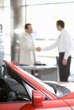 Photo: Salesman shaking hands with male customer in car showroom, focus on red convertible in foreground Salesman shaking hands with male customer in car showroom, focus on red convertible in foreground