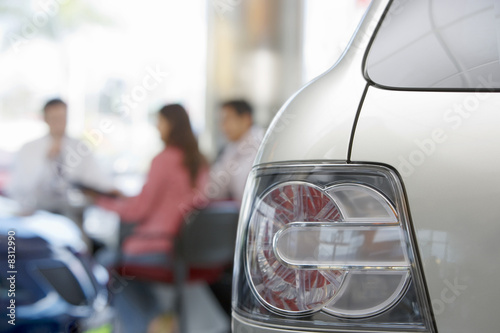 Photo: Salesman and couple sitting at desk in car showroom, focus on tail light in foreground Salesman and couple sitting at desk in car showroom, focus on tail light in foreground