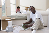 Photo: Couple moving house, woman with lamp, focus on man unpacking glassware in living room, smiling, portrait Couple moving house, woman with lamp, focus on man unpacking glassware in living room, smiling, portrait