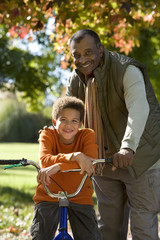 Photo: Father standing with son (7-9) in autumn park, boy sitting on bicycle, smiling, portrait Father standing with son (7-9) in autumn park, boy sitting on bicycle, smiling, portrait