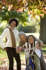 Photo: Girl (7-9) sitting on bicycle beside mother and grandmother in autumn park, senior woman embracing granddaughter, portrait Girl (7-9) sitting on bicycle beside mother and grandmother in autumn park, senior woman embracing granddaughter, portrait