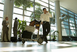 Photo: Businessman, with luggage trolley, standing in airport terminal, using mobile phone (surface level) Businessman, with luggage trolley, standing in airport terminal, using mobile phone (surface level)