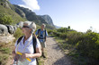 Photo: Mature couple, with rucksacks and hiking poles, hiking on mountain trail, woman leading, front view Mature couple, with rucksacks and hiking poles, hiking on mountain trail, woman leading, front view