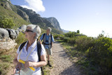 Photo: Mature couple, with rucksacks and hiking poles, hiking on mountain trail, woman leading, front view Mature couple, with rucksacks and hiking poles, hiking on mountain trail, woman leading, front view