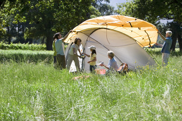 Photo: Multi-generational family assembling orange tent on camping trip in woodland clearing, side view Multi-generational family assembling orange tent on camping trip in woodland clearing, side view
