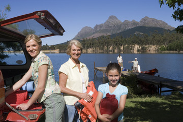 Photo: Girl (8-10) unloading life jackets from SUV with mother and grandmother, rest of family on lake jetty beside boat Girl (8-10) unloading life jackets from SUV with mother and grandmother, rest of family on lake jetty beside boat