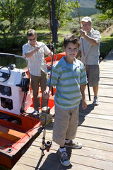 Photo: Boy (8-10) standing on lake jetty with fishing rod, father and grandfather loading motorboat in background Boy (8-10) standing on lake jetty with fishing rod, father and grandfather loading motorboat in background