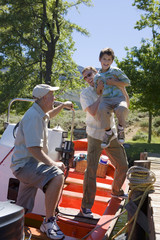 Photo: Father lifting son (8-10) onto motorboat moored at lake jetty, grandfather looking on, smiling Father lifting son (8-10) onto motorboat moored at lake jetty, grandfather looking on, smiling
