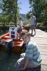 Photo: Boy (8-10) sitting on lake jetty, smiling, side view, portrait, father and grandfather loading fishing rods into motorboat Boy (8-10) sitting on lake jetty, smiling, side view, portrait, father and grandfather loading fishing rods into motorboat