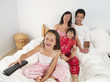 Photo: Family sitting on double bed at home, watching television, girl (5-7) using remote control, laughing Family sitting on double bed at home, watching television, girl (5-7) using remote control, laughing