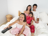 Photo: Family sitting on double bed at home, watching television, girl (5-7) using remote control, laughing Family sitting on double bed at home, watching television, girl (5-7) using remote control, laughing