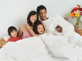 Photo: Family sitting in double bed at home, watching television, man using remote control Family sitting in double bed at home, watching television, man using remote control