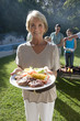 Photo: Three generation family standing beside barbecue grill in garden, focus on senior woman holding plate of food in foreground, smiling, portrait Three generation family standing beside barbecue grill in garden, focus on senior woman holding plate of food in foreground, smiling, portrait