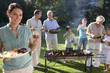 Foto: Three generation family standing beside barbecue grill in garden, focus on woman holding plate of food and glass of wine in foreground, smiling, portrait Three generation family standing beside barbecue grill in garden, focus on woman holding plate of food and glass of wine in foreground, smiling, portrait