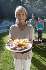Photo: Three generation family standing beside barbecue grill in garden, focus on senior woman holding plate of food in foreground, smiling, portrait Three generation family standing beside barbecue grill in garden, focus on senior woman holding plate of food in foreground, smiling, portrait