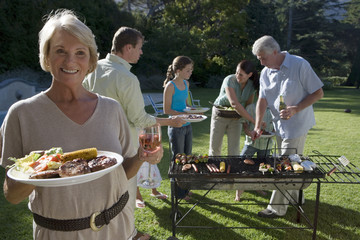 Photo: Three generation family standing beside barbecue grill in garden, focus on senior woman holding plate of food in foreground, smiling, portrait Three generation family standing beside barbecue grill in garden, focus on senior woman holding plate of food in foreground, smiling, portrait