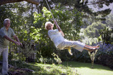 Photo: Senior woman swinging on garden rope swing, man pushing woman, smiling, side view, portrait Senior woman swinging on garden rope swing, man pushing woman, smiling, side view, portrait