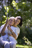 Photo: Senior woman swinging on garden rope swing, adult daughter embracing her, smiling, front view, portrait Senior woman swinging on garden rope swing, adult daughter embracing her, smiling, front view, portrait