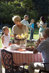 Photo: Three generation family having barbecue in summer garden, girl (8-10) standing beside grandmother at table, woman serving corn cobs, smiling Three generation family having barbecue in summer garden, girl (8-10) standing beside grandmother at table, woman serving corn cobs, smiling