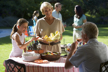 Photo: Three generation family having barbecue in summer garden, girl (8-10) standing beside grandmother at table, woman serving corn cobs, smiling Three generation family having barbecue in summer garden, girl (8-10) standing beside grandmother at table, woman serving corn cobs, smiling