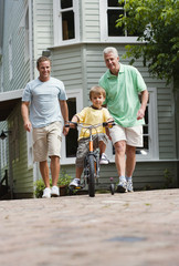 Photo: Boy (4-6) riding bicycle with stabilisers on pavement, father and grandfather looking on, smiling, front view, portrait (surface level) Boy (4-6) riding bicycle with stabilisers on pavement, father and grandfather looking on, smiling, front view, portrait (surface level)