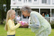 Photo: Girl (4-6) presenting birthday gift to grandmother in summer garden, smiling, profile, family members on house veranda in background Girl (4-6) presenting birthday gift to grandmother in summer garden, smiling, profile, family members on house veranda in background