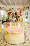 Photo: Three generation family celebrating birthday at home, smiling, portrait, birthday cake on table in foreground Three generation family celebrating birthday at home, smiling, portrait, birthday cake on table in foreground