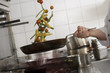 Photo: Male chef tossing stir fry vegetables in frying pan over gas hob in commercial kitchen, side view, mid-section (tilt) Male chef tossing stir fry vegetables in frying pan over gas hob in commercial kitchen, side view, mid-section (tilt)