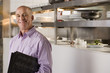 Photo: Male restaurant manager standing in commercial kitchen, carrying menus, smiling, side view, portrait Male restaurant manager standing in commercial kitchen, carrying menus, smiling, side view, portrait