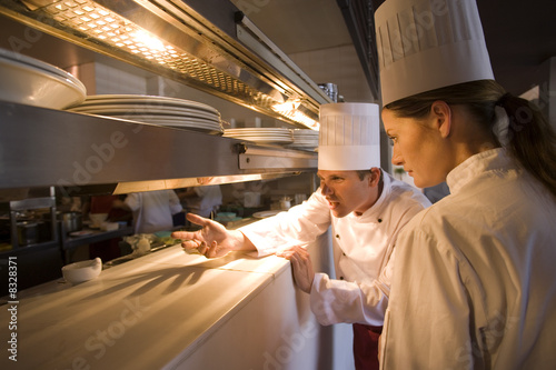 Photo: Male and female chefs standing at order counter in commercial kitchen, agitated male chef gesturing to restaurant staff Male and female chefs standing at order counter in commercial kitchen, agitated male chef gesturing to restaurant staff