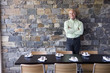 Photo: Mature male restaurant owner standing beside table, leaning against stone wall, smiling, front view, portrait Mature male restaurant owner standing beside table, leaning against stone wall, smiling, front view, portrait