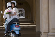 Photo: Couple riding on blue motor scooter near colonnade, woman holding bouquet of flowers, looking over shoulder, smiling, rear view, portrait Couple riding on blue motor scooter near colonnade, woman holding bouquet of flowers, looking over shoulder, smiling, rear view, portrait