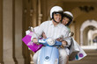 Photo: Senior couple riding on motor scooter near colonnade, woman holding shopping bags, smiling, front view Senior couple riding on motor scooter near colonnade, woman holding shopping bags, smiling, front view