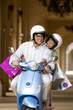 Photo: Senior couple riding on motor scooter near colonnade, woman holding shopping bags, smiling, front view Senior couple riding on motor scooter near colonnade, woman holding shopping bags, smiling, front view